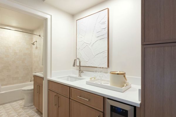 Modern kitchenette with wood cabinets, white countertop, abstract wall art, and a small fridge; a bathroom is visible in the background.