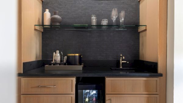 A small bar setup with shelves holding glassware and vases, a countertop with a sink, and a mini-fridge below.