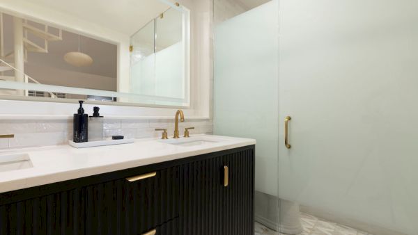 A modern bathroom with a dark vanity, gold fixtures, a large mirror, and a frosted glass shower door on a patterned white floor.
