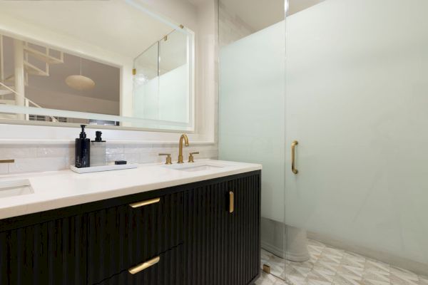 A modern bathroom with a dark vanity, gold fixtures, a large mirror, and a frosted glass shower door on a patterned white floor.