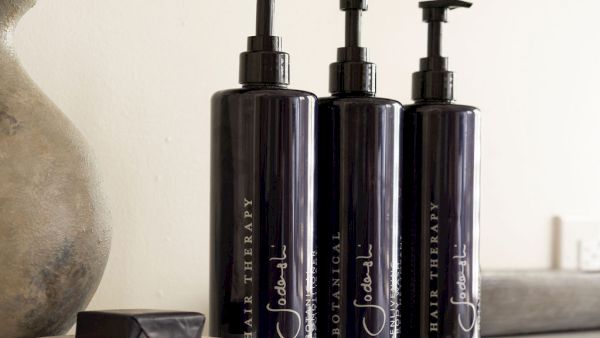 The image shows three dark pump bottles labeled "Hair Therapy" and "Botanical," alongside a small dish with a dark bar, on a light shelf.