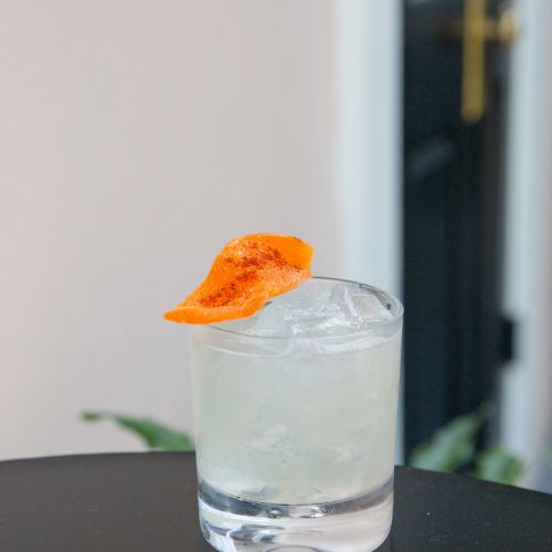 A cocktail in a glass with ice, garnished with an orange slice, sits on a black table.
