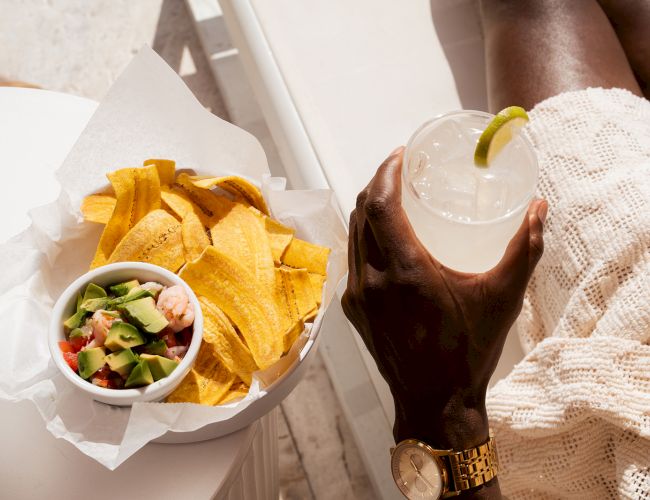 A person is holding a drink with a lime slice, next to a bowl of chips and salsa on a table, while sitting in a relaxed setting.