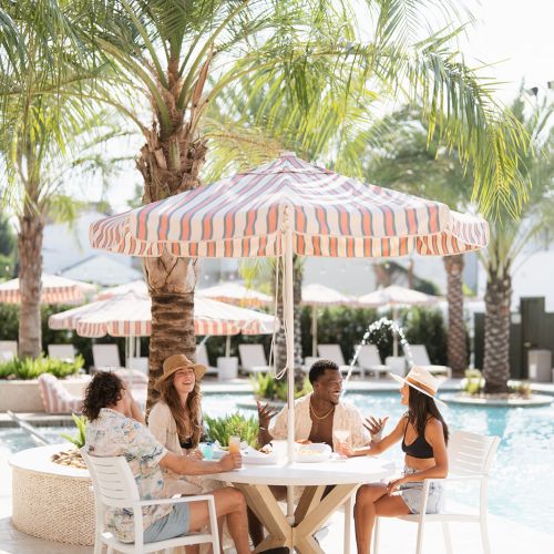 A group of people sitting and talking around a table with a striped umbrella by a pool, surrounded by palm trees.