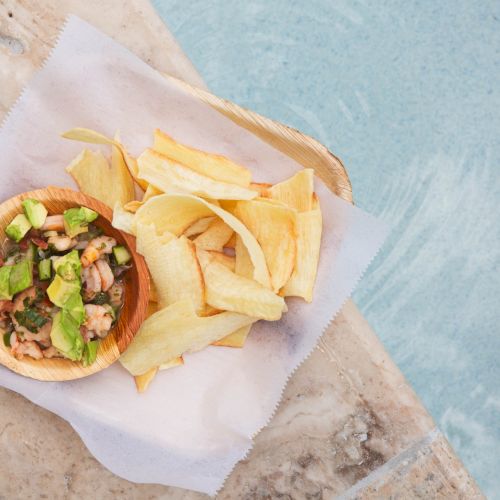 The image shows a bowl of food with avocado and chips on a napkin, placed by a poolside.