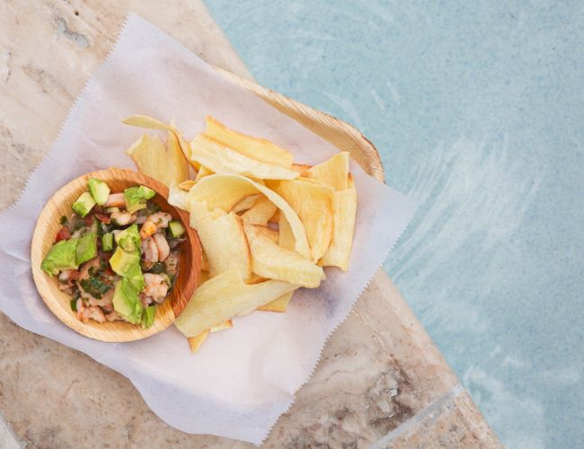 The image shows a bowl of food with avocado and chips on a napkin, placed by a poolside.