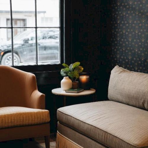 The image shows a cozy corner with a beige armchair, a round table with a plant and candle, next to a window with a view of a parked car.