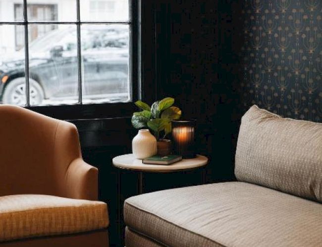 The image shows a cozy corner with a beige armchair, a round table with a plant and candle, next to a window with a view of a parked car.