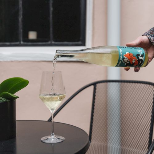 A person pours white wine into a glass on a black table beside a potted plant and a metal chair with a window in the background.