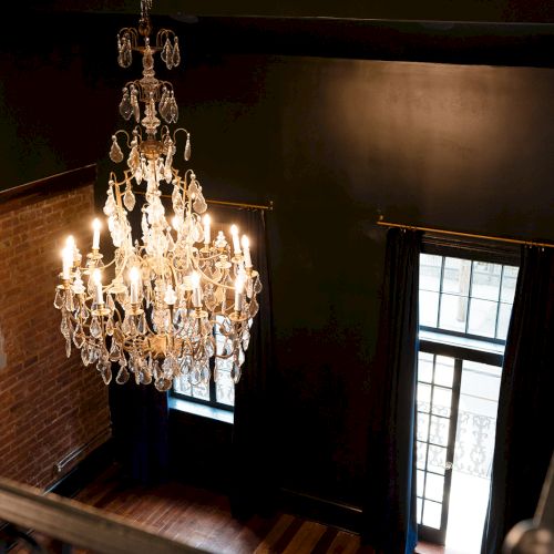 A grand crystal chandelier hangs in a brick-walled room with lit candles and curtained windows.
