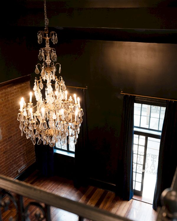 A grand crystal chandelier hangs in a brick-walled room with lit candles and curtained windows.