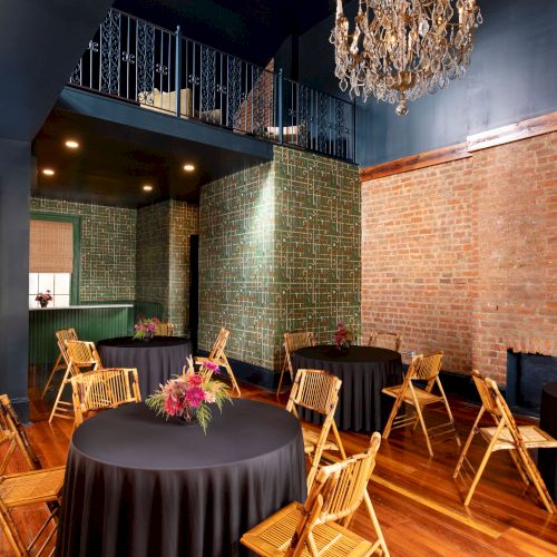 A stylish room with round tables, black tablecloths, wooden chairs, brick walls, and a chandelier hanging from a high ceiling.