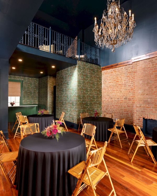 A stylish room with round tables, black tablecloths, wooden chairs, brick walls, and a chandelier hanging from a high ceiling.
