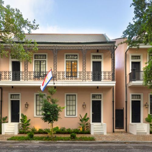A row of picturesque terraced houses with balconies, flags, and lush greenery lining the sidewalk, under a canopy of trees.