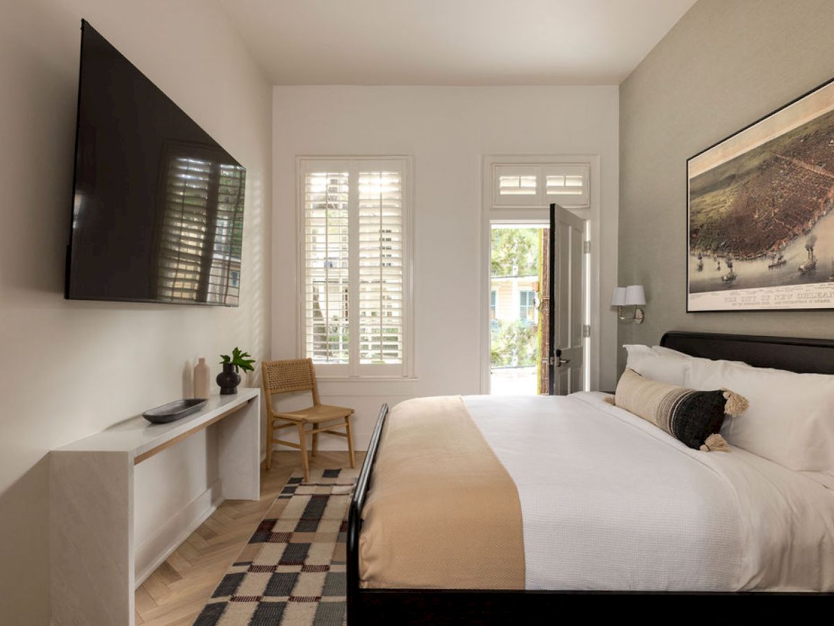 A modern bedroom with a large bed, wall-mounted TV, artwork, and a console table. Light streams through shutters on the window.