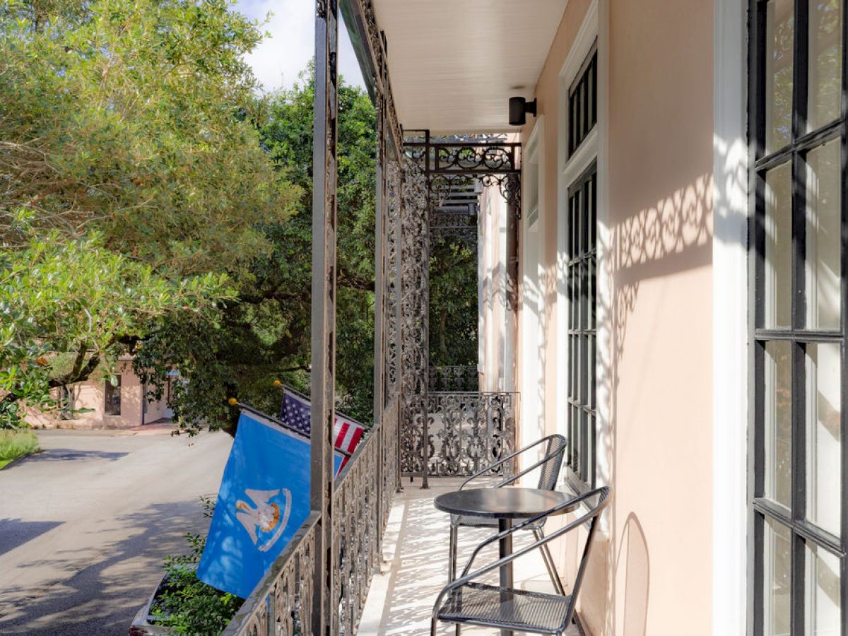 A balcony with iron railings overlooks a leafy street, featuring two chairs, a small table, and two flags.