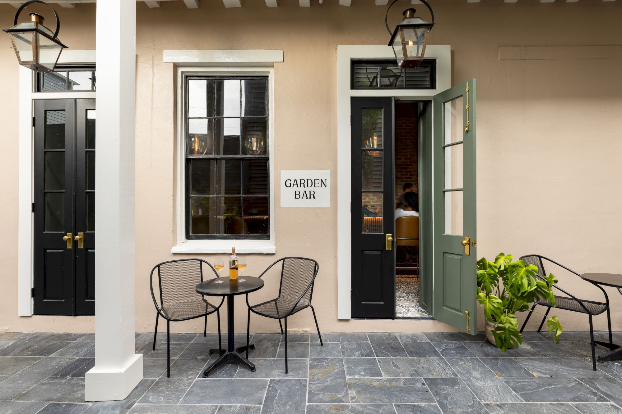 A cozy outdoor patio with two round tables, chairs, and a sign reading "Garden Bar" near open doors, set on a stone tiled floor.