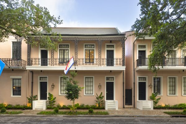 The image shows a row of charming townhouses with balconies and trees, featuring American and other flags in front, under a blue sky.
