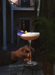 A hand holds a cocktail glass with a frothy drink, garnished with purple and yellow edible flowers, against a dimly lit outdoor setting.