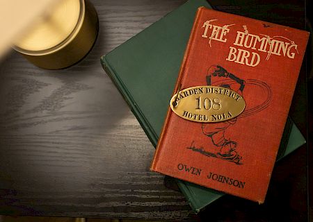 A candle, a red book titled "The Humming Bird," and a green book are on a dark wooden table. A hotel emblem is on the red book.