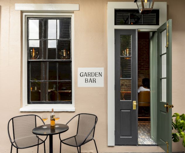 A cozy outdoor space with two chairs, a table with a bottle, and a "Garden Bar" sign beside a door.