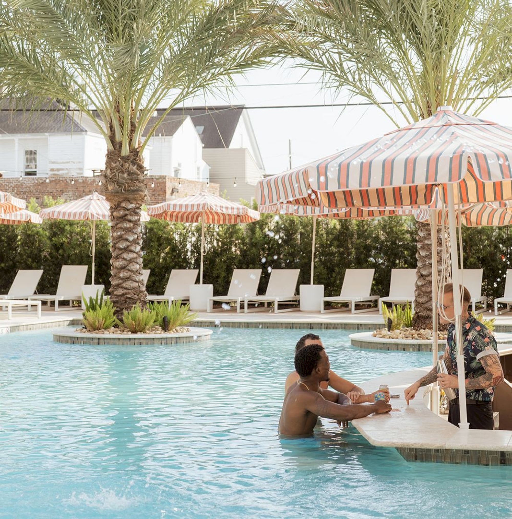 A pool scene with two people at a swim-up bar under striped umbrellas, surrounded by palm trees and lounge chairs in the background.