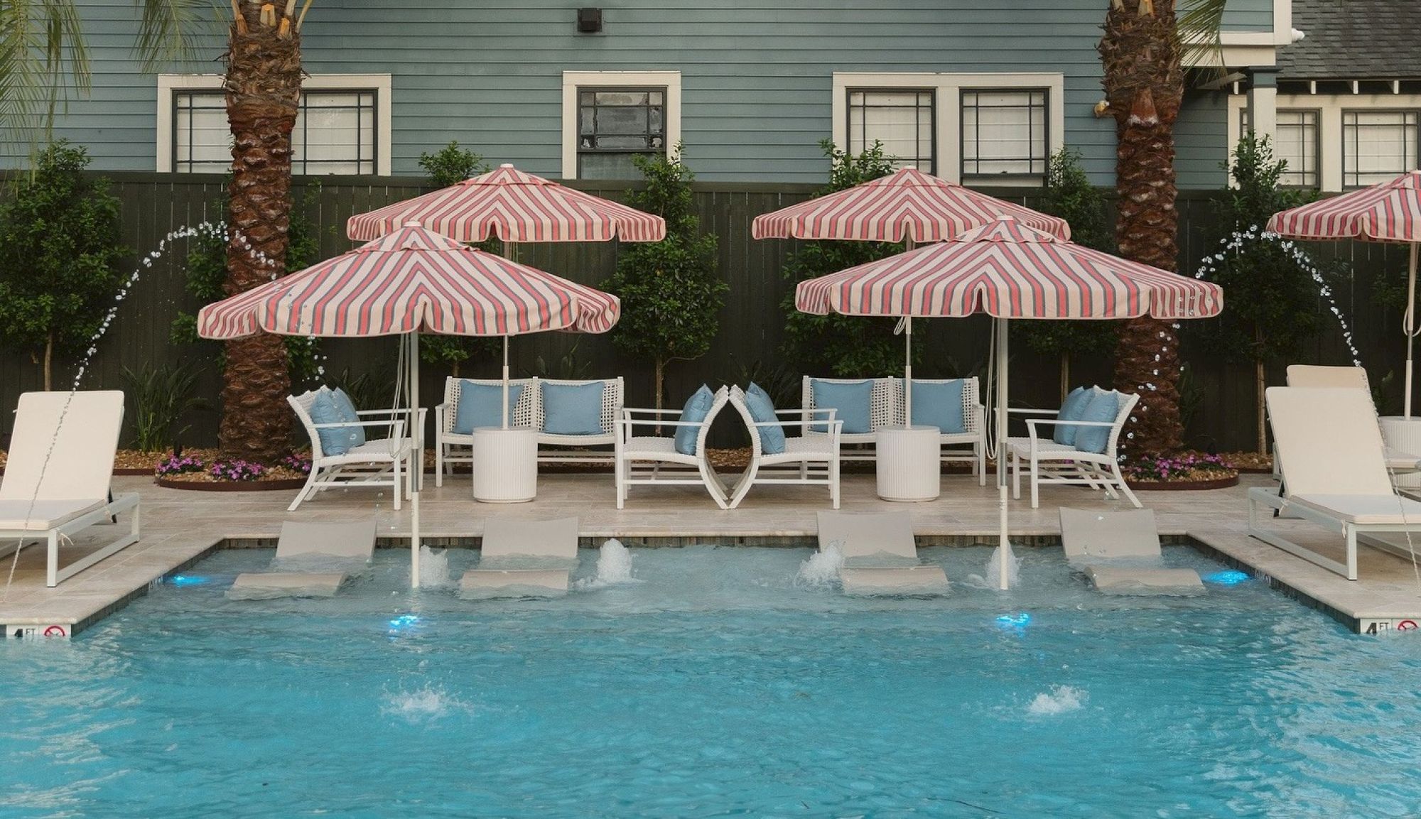 Poolside scene with loungers, red-striped umbrellas, and water jets; framed by a green fence and palm trees, creating a relaxing vibe.
