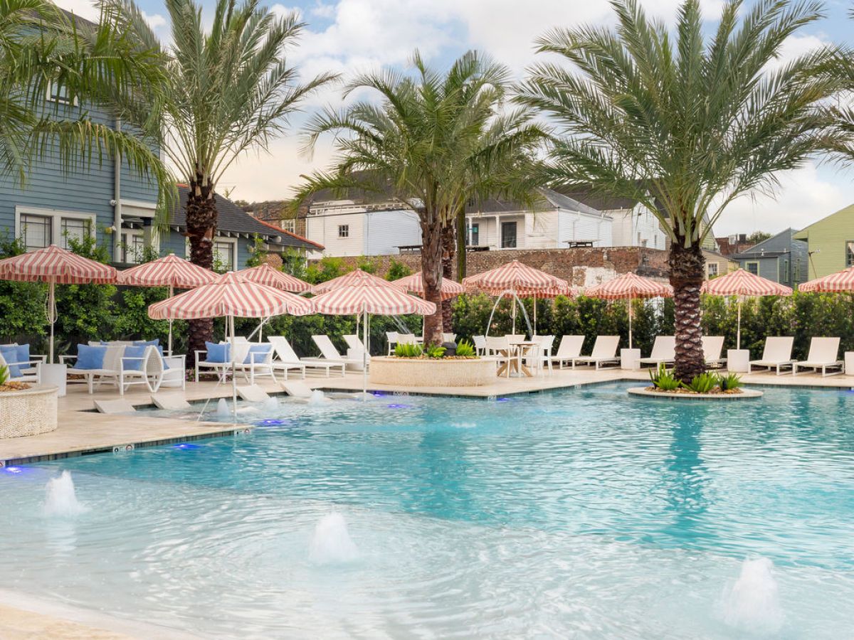 A serene swimming pool surrounded by palm trees, sun loungers, and pink umbrellas under a blue sky with fluffy clouds.