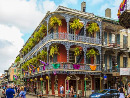 A street view of a historic building with ornate balconies, colorful flags, and greenery; pedestrians and a parked car are visible.