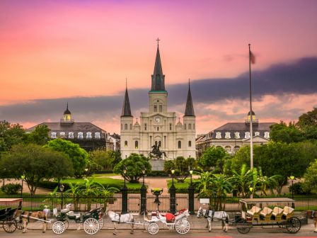 A historic cathedral with three spires stands against a colorful sky, with carriages lined up in front, and lush gardens surrounding it.