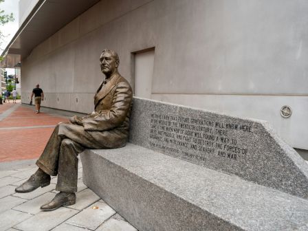 A bronze statue of a seated man on a bench with engraved text, located along a sidewalk near a building.