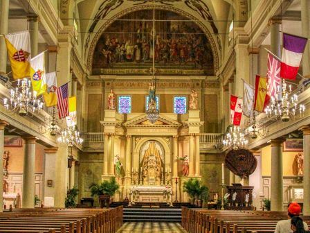 A church interior with ornate ceilings, flags, chandeliers, and a checkered floor leading to an altar at the end of the aisle.