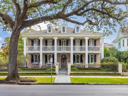 A grand, two-story house with columns and balconies, surrounded by greenery and a large tree in the foreground.