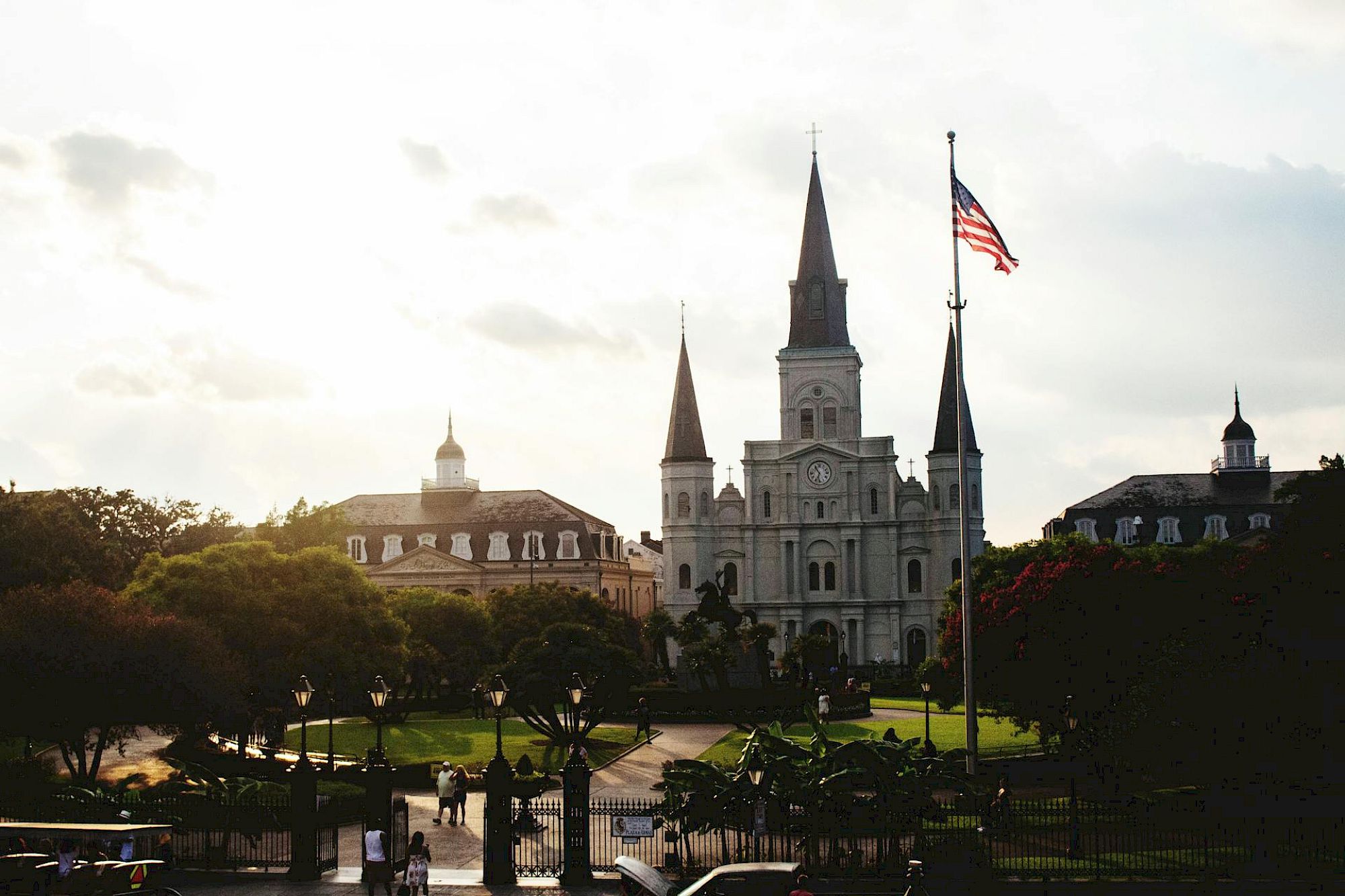The image shows a historic cathedral with three spires, an American flag, and a park in the foreground under a cloudy sky.