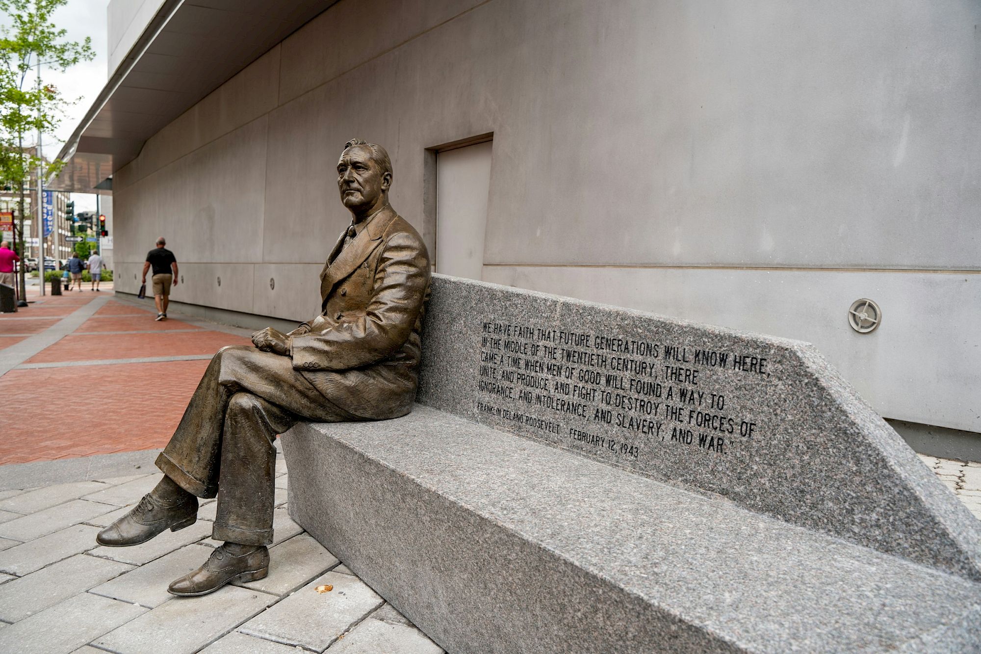 A bronze statue of a seated man on a bench, with engraved text, is in an urban street setting.
