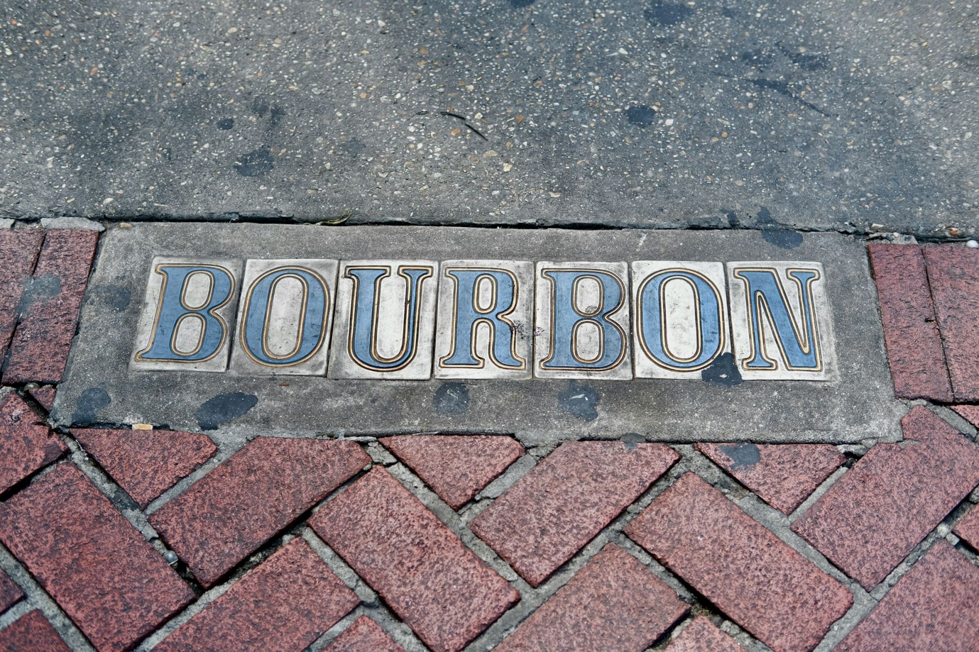 The image shows the word "Bourbon" written in decorative tiles set into a sidewalk with red brick paving.