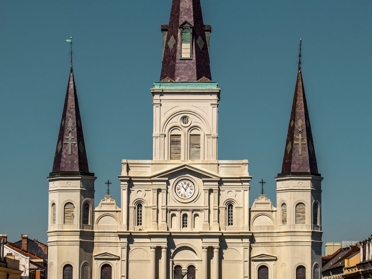 The image shows a large, historic cathedral with three spires, surrounded by a park, under a clear blue sky.