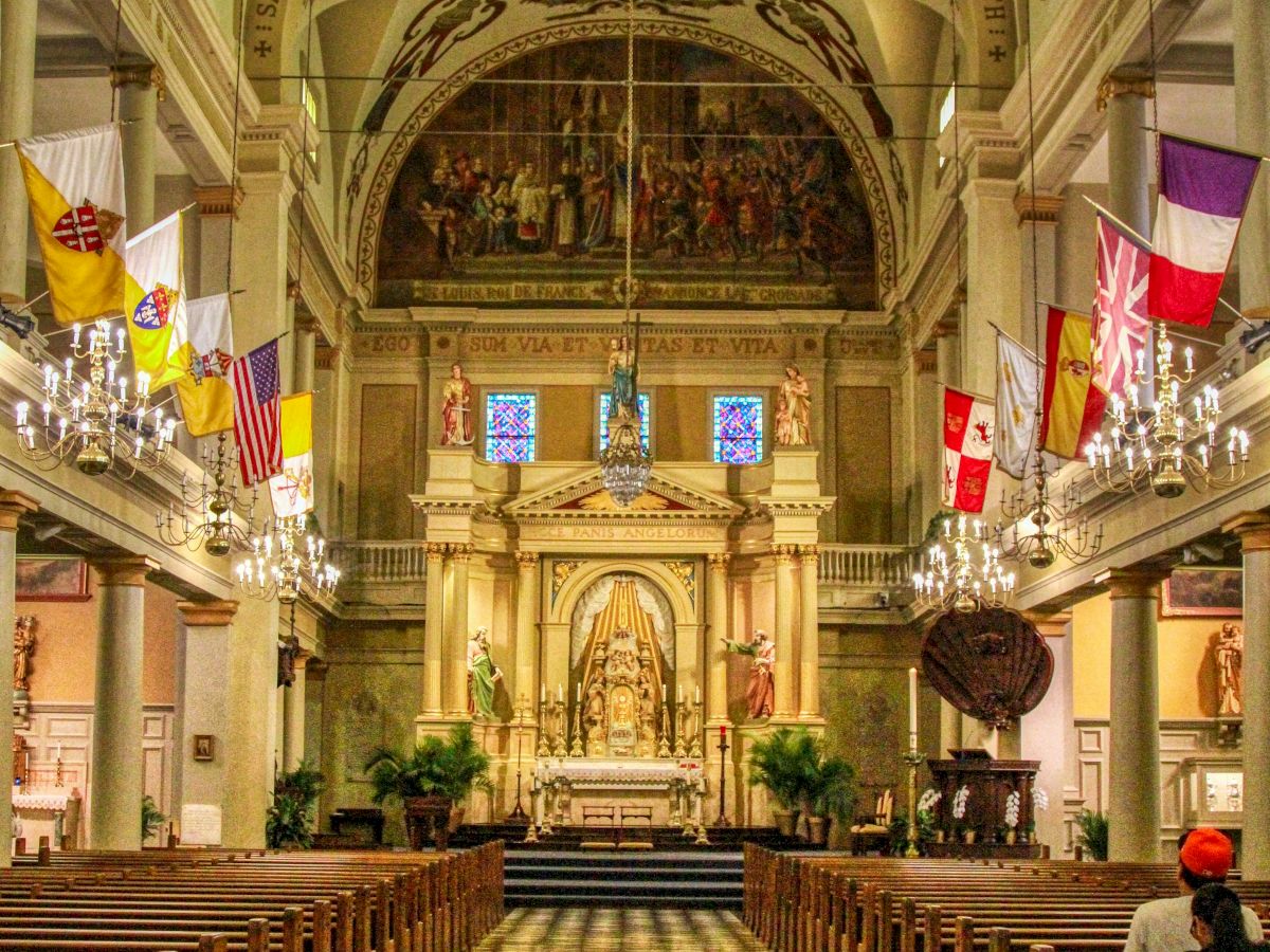 An ornate church interior with checkered floor, chandeliers, flags, and a detailed ceiling leading to an altar.
