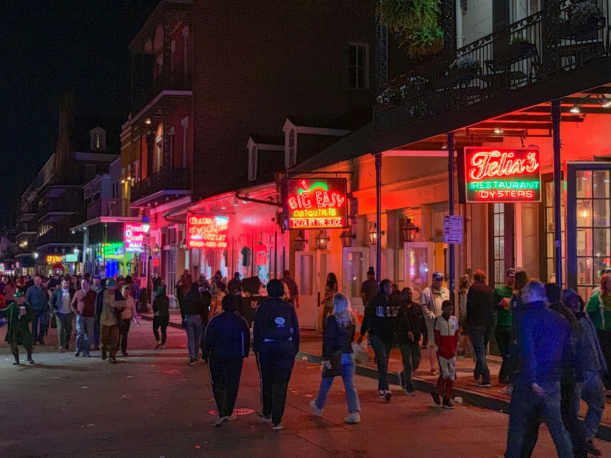 A lively street scene at night with neon signs and people walking by restaurants and bars, creating a bustling atmosphere.
