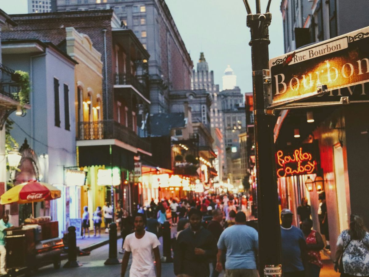 A bustling city street features a crowd of people, colorful buildings, and signs with "Bourbon" in an evening urban scene.