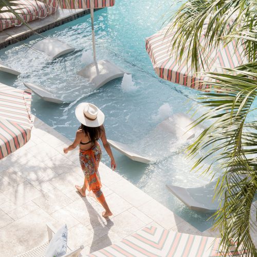 A person in a sun hat walks beside a pool with striped umbrellas and lounge chairs, surrounded by tropical plants.