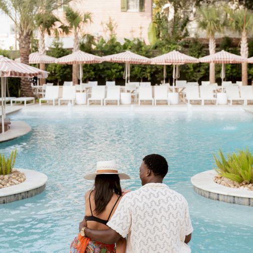 A couple sits by a pool, surrounded by palm trees and lounge chairs with umbrellas, enjoying a peaceful and sunny day together.