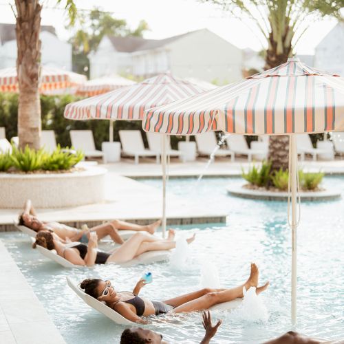 People are relaxing on lounge chairs in a pool under striped umbrellas, with palm trees and sunny skies around them.