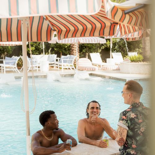 Three people enjoy drinks at a poolside bar, one standing and two in the pool, under a striped umbrella on a sunny day.