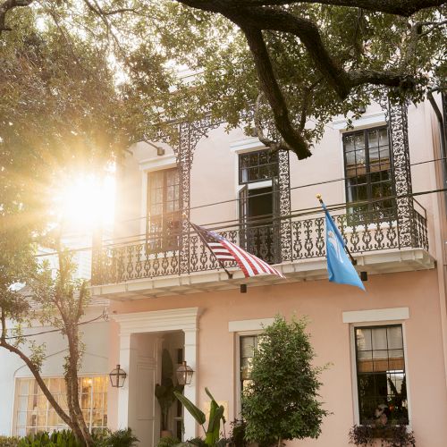 A charming two-story house with decorative ironwork and two flags. The sunlight peeks through the trees, creating a warm atmosphere.