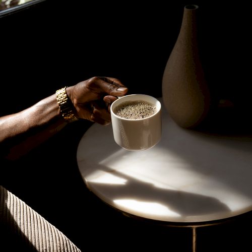 A person holding a coffee cup over a small table with a vase, in a dimly lit setting.