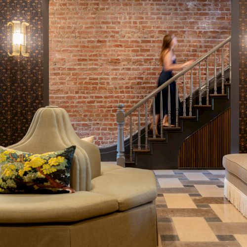 A stylish room with an octagonal sofa, patterned wallpaper, exposed brick wall, and a person on the stairs. A cushion adds color.