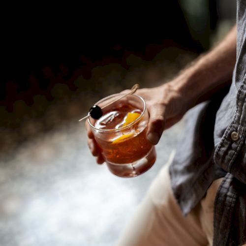 A person is holding a glass of cocktail garnished with an orange slice and cherry, dressed in casual clothing.