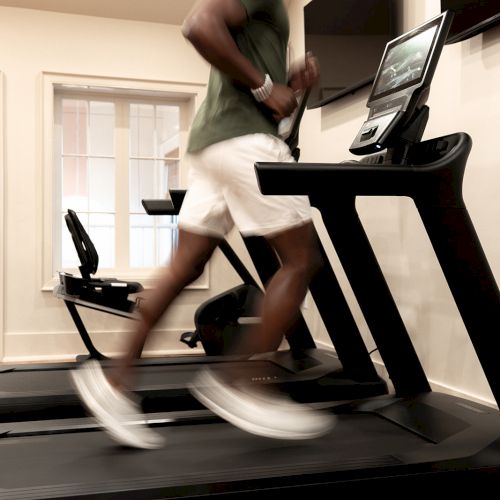 A person is running on a treadmill in a well-lit room with wooden floors and a large window.