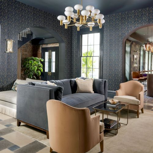 A stylish living room with a dark blue ceiling, modern chandelier, grey sofa, beige chairs, patterned rug, and a plant by the window.